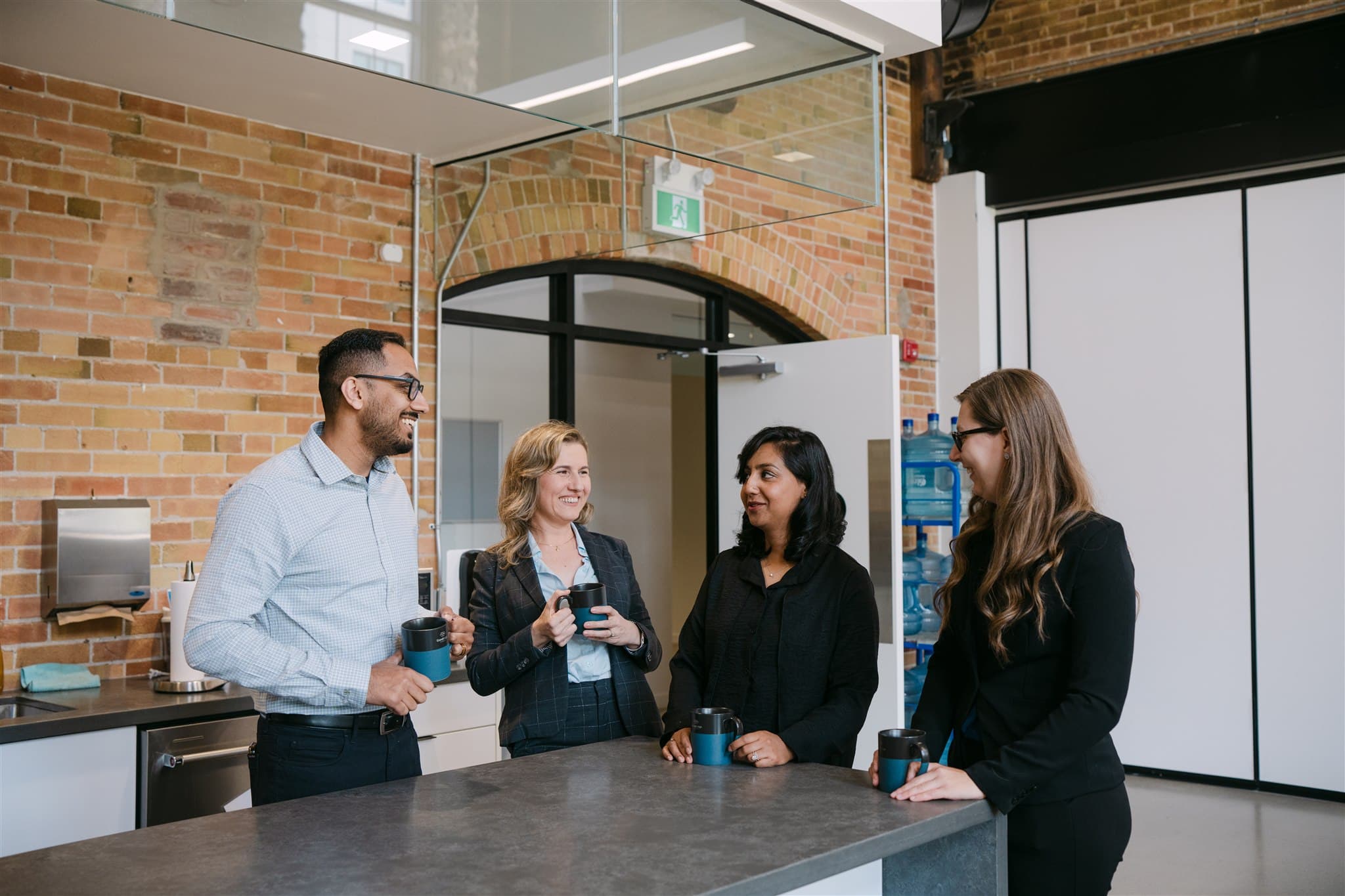 Four people in business attire stand in an office kitchen, holding mugs and talking by a counter.