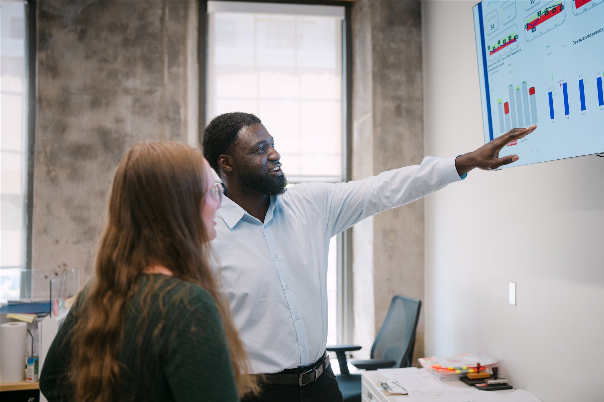 A man points at a data chart on a screen while discussing it with a woman in an office setting.