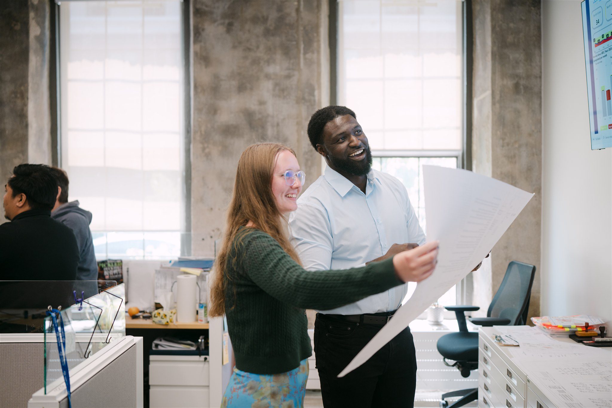 Two employees comparing a blueprint to a computer screen on the wall.