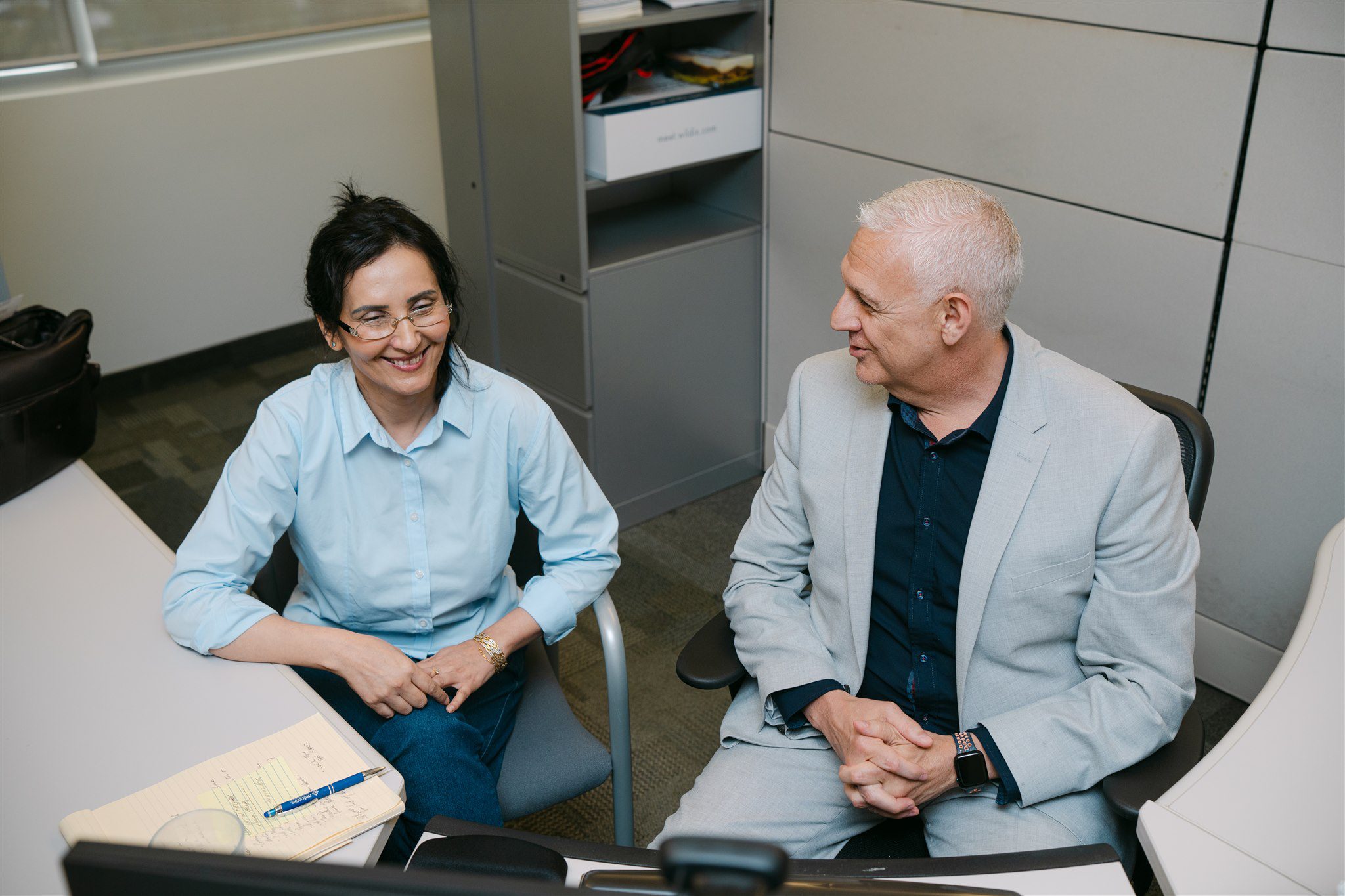 Two colleagues sit at a desk in an office, talking and smiling, with papers and a pen on the table.