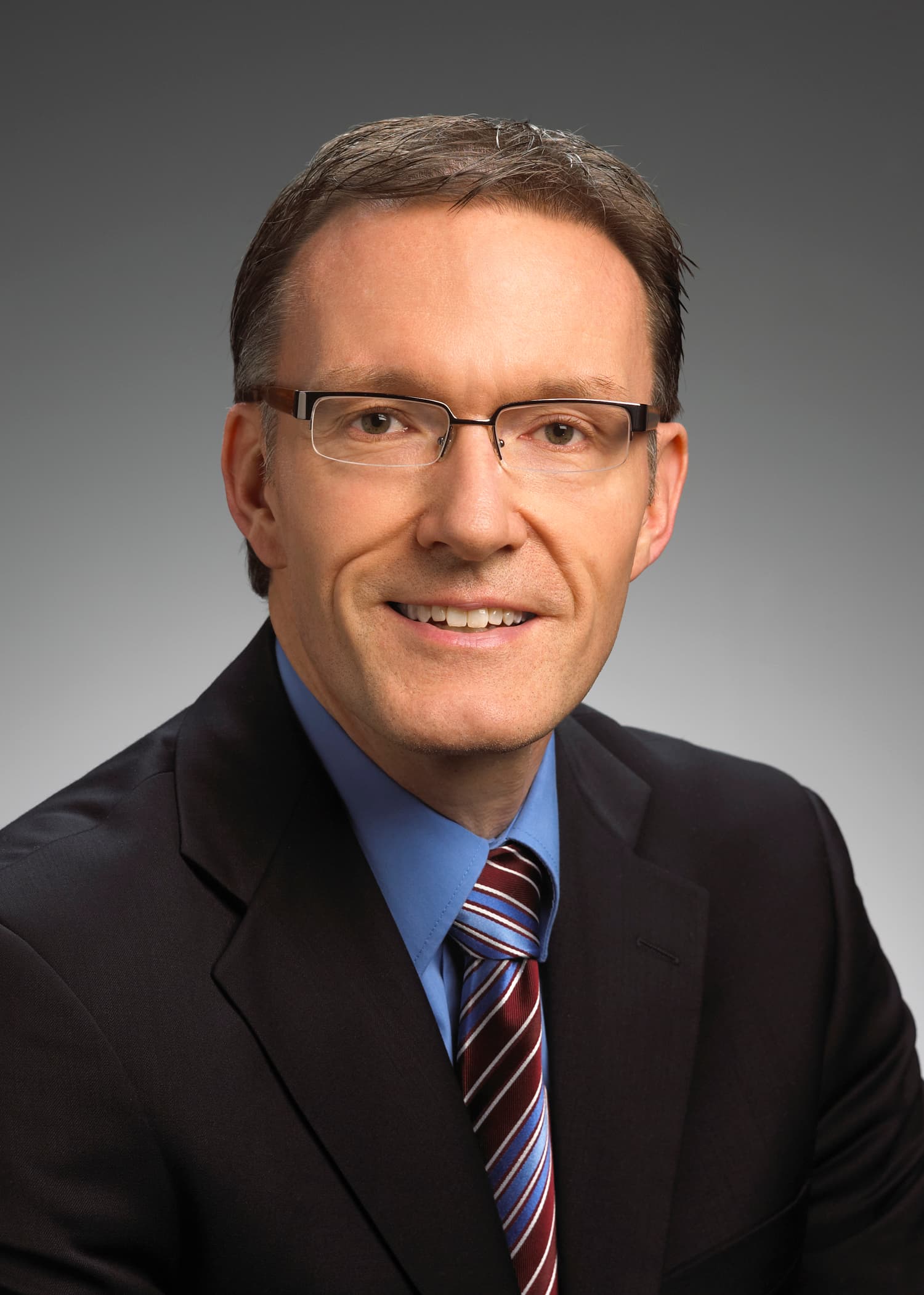 Man in a suit and tie with glasses, short brown hair, and a neutral background, facing the camera and smiling.